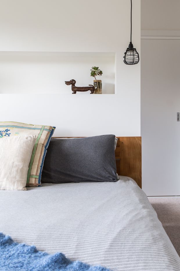 Bright, calm bedroom interior with a neatly made bed, layered neutral and blue bedding, a timber headboard, white walls, a recessed display niche with decorative objects and a plant, and a black wire pendant light. A minimal sliding or hinged door is visible to the right.