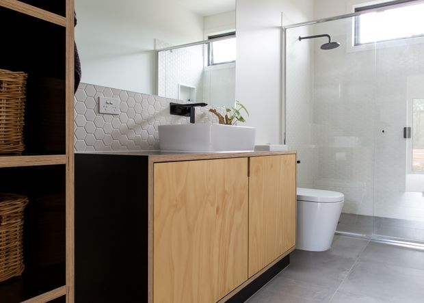 Bright contemporary bathroom featuring a timber vanity with a white vessel basin, geometric hexagonal tile splashback, black wall-mounted tapware, open shelving with woven baskets, and a frameless glass shower enclosure. Grey floor tiles and high windows contribute to a clean, airy feel.