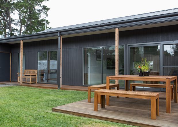 Single-storey dark-clad house with a low-pitched roof, large glazed sliding doors, and a timber-framed covered deck. The deck includes a simple timber dining table and benches, opening onto a neatly landscaped lawn. Tall trees are visible in the background.