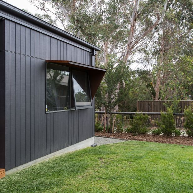 Exterior view of a compact contemporary building with dark vertical cladding, a projecting angled window awning, and a simple low-pitched roof. The structure sits beside a landscaped lawn and mature eucalyptus trees, with fencing visible in the background.