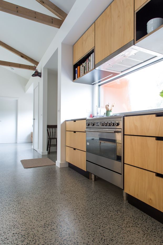 Bright contemporary kitchen with a stainless-steel freestanding oven and cooktop, extensive light timber cabinetry with black recessed pulls, open shelving, and a large window over the benchtop. The kitchen opens to a hallway with white walls, exposed timber ceiling beams, and a speckled polished concrete or terrazzo-like floor.