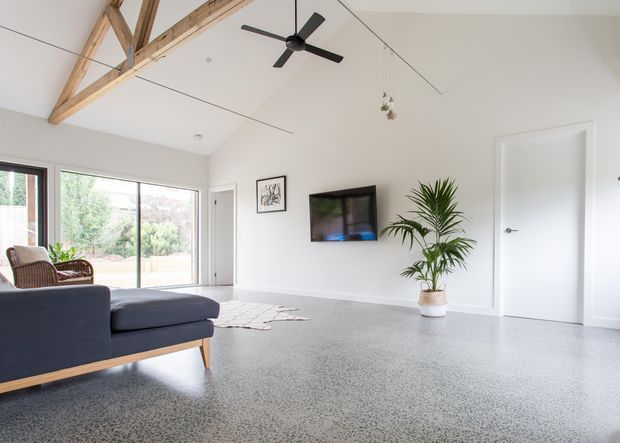Bright, open-plan living room with polished speckled concrete flooring, white walls, exposed timber roof beams, and large sliding glass doors overlooking greenery. A dark sectional sofa, wall-mounted television, indoor palm, ceiling fan, and minimal furnishings create a clean contemporary interior.