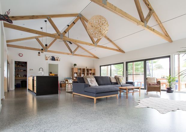 Bright open-plan living area with a dark kitchen island, large sectional sofa, dining area, extensive glazing, and a dramatic pitched ceiling with exposed timber trusses and beams. The polished speckled floor and white walls create a clean, airy interior, while a woven pendant light and indoor plants add warmth.