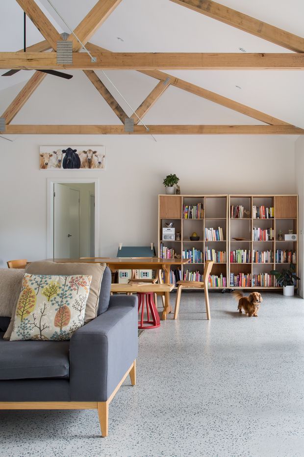 Bright open-plan living and study space with a large grey sofa in the foreground, a timber desk and chairs, extensive built-in timber bookshelves, and a small dog. The room has a high pitched ceiling with exposed timber post-and-beam rafters, white walls, and a light terrazzo-style floor.