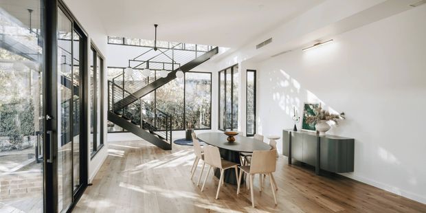 Bright contemporary open-plan dining area with a black-framed glazed stairwell and floating staircase, light timber flooring, white walls, and generous floor-to-ceiling windows. A round dining table with upholstered chairs sits beside a dark built-in sideboard, with abundant natural light and views of surrounding trees.