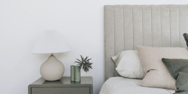 A close-up of a softly styled bedroom corner featuring a bed with a tall vertically channelled light-grey upholstered headboard, layered white and patterned cushions, and a grey bedside table. A rounded ceramic lamp and small potted plant sit on the table against a plain light wall.