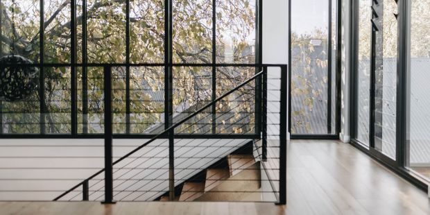 A bright contemporary interior landing beside a stairwell, featuring floor-to-ceiling black-framed glazing, pale timber flooring, white walls, and a minimalist black metal stair balustrade with horizontal wire infill. Trees and neighbouring structures are visible through the windows.