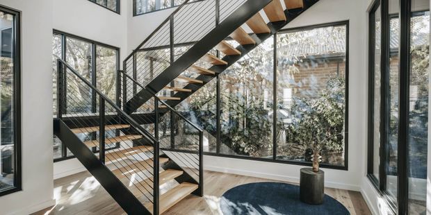 Bright contemporary stairway with an open-riser timber staircase, black metal stringers and slim wire balustrades. Floor-to-ceiling glazing surrounds the stair, bringing in abundant daylight and views of trees, while pale timber flooring, white walls and a dark round rug create a clean, modern interior.
