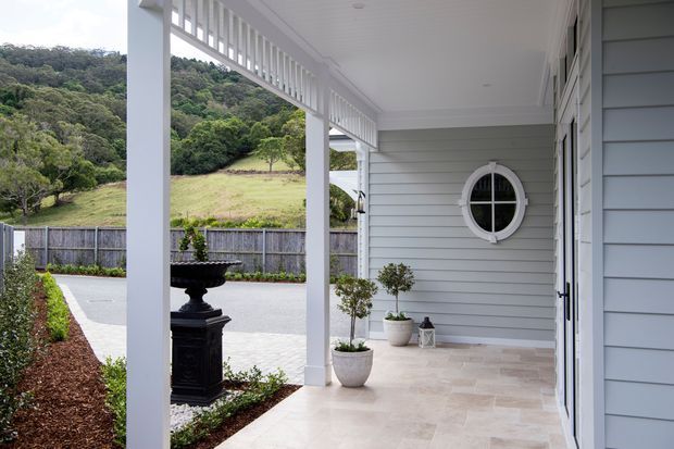 A covered front porch and entryway with white-painted posts, balustrade detailing, pale horizontal weatherboard cladding, and a round divided-pane window. The porch has a light stone-tile floor, potted topiary, a black decorative urn, and a dark entry door, overlooking a fenced landscaped rural setting.