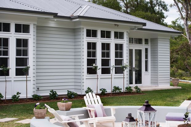 Exterior view of a light-grey weatherboard house with a low-pitched metal roof, white-trimmed multi-pane windows and glazed French-style doors. A furnished paved outdoor area with Adirondack chairs and lanterns sits in the foreground, with landscaped planting around the building.