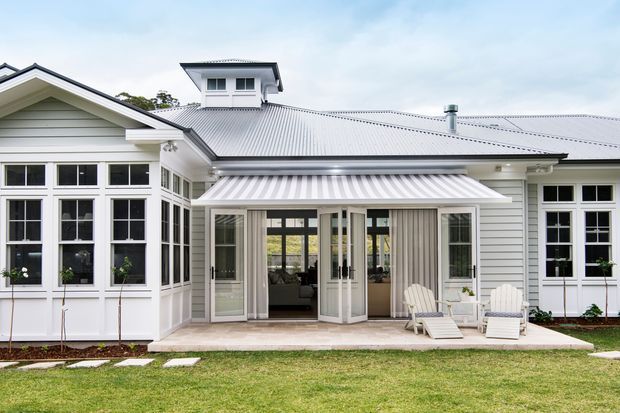 Exterior rear view of a light-painted, single-storey home with a broad corrugated metal roof, central raised roof monitor, gabled window bay, and large multi-pane windows. A covered veranda or terrace with folding glass doors opens to a paved outdoor seating area and lawn. The facade features horizontal weatherboards and white trim in a bright, traditional residential style.