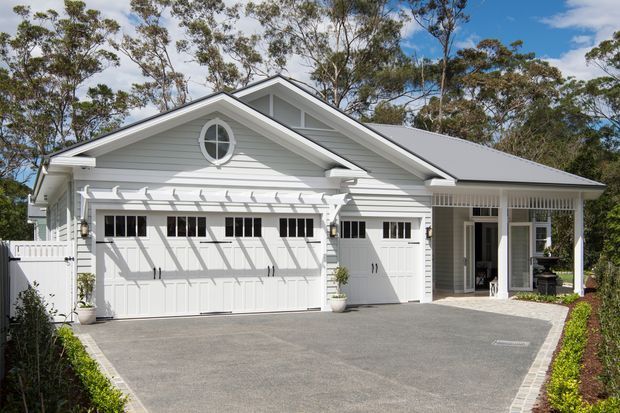 A bright white, gabled residential garage and entry facade set among trees. The building features horizontal weatherboard cladding, a large double garage door, a separate single garage or service door, a covered porch with slender columns, a circular attic window, and a broad paved driveway.