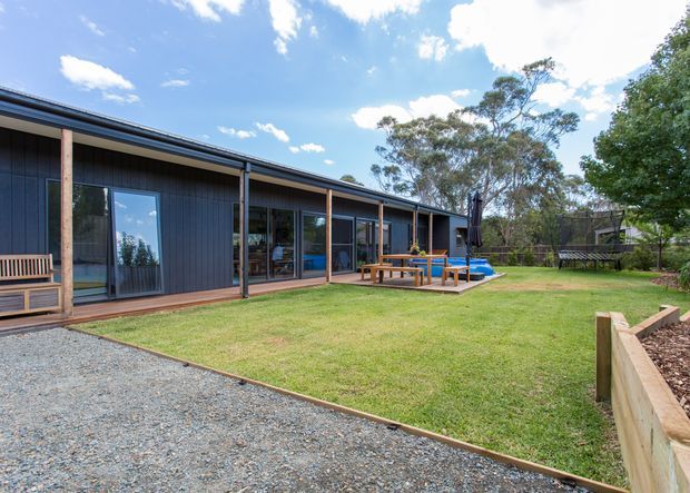 Single-storey contemporary dark-clad residence set within a landscaped suburban or bush-like garden. The long low facade has a shallow pitched roof, extensive glazed sliding doors, a timber deck, outdoor dining area, lawn, gravel path, raised garden edging and a small visible pool or water feature. Mature trees surround the property.