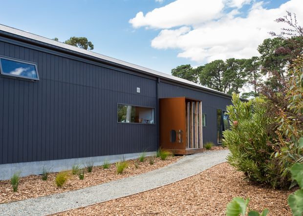 Single-storey contemporary building exterior with dark vertical cladding, a shallow pitched roof, and a warm timber entry volume. A curved gravel walkway leads through landscaped garden beds to the recessed glazed entrance.
