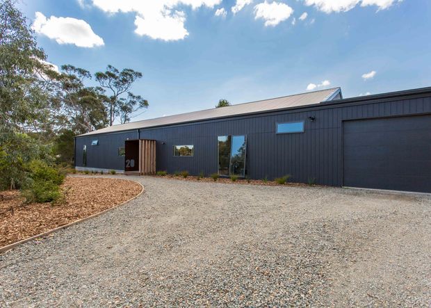 Single-storey contemporary dark-clad building viewed from a broad gravel driveway. The facade features vertical dark cladding, a large integrated garage door, timber entrance screening, narrow horizontal windows, and a pitched metal roof, set within a lightly landscaped bush-like setting.