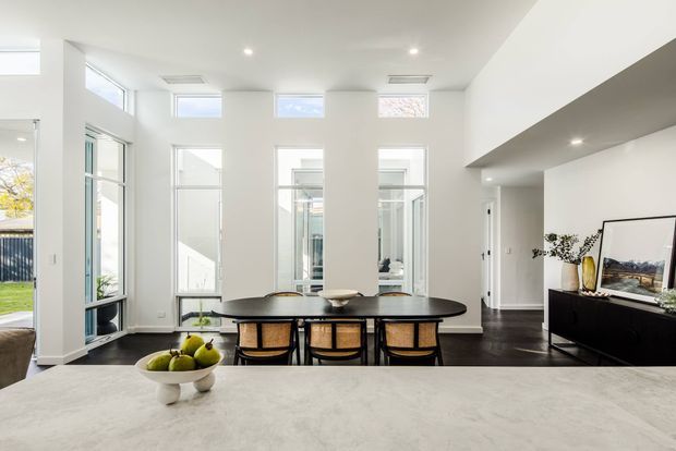 Bright contemporary open-plan dining and kitchen interior with a long dark oval dining table and woven-back chairs positioned beside extensive white-framed glazing. A pale stone kitchen island occupies the foreground, while a dark media console and artwork sit along the right wall. The room features a white double-height ceiling volume, recessed downlights, tall windows, and dark timber flooring.