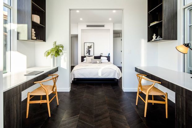 Bright, symmetrical bedroom with a central sleeping area set behind an open threshold and flanked by two compact workstations. The room features dark herringbone timber flooring, white walls and ceiling, integrated dark timber cabinetry and desks, pale upholstered bedding, and matching light timber chairs. Tall windows provide abundant natural light, while recessed shelving and a concealed central air-conditioning vent contribute to the clean, refined design.