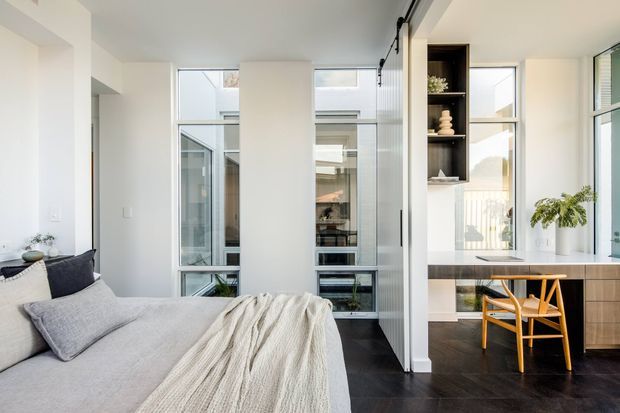 Bright contemporary bedroom with a neatly made bed in soft grey and cream textiles, dark timber flooring, full-height glazing, and a compact built-in study nook with timber cabinetry, open shelving, and a wooden chair. White walls and abundant natural light create an airy, refined atmosphere.