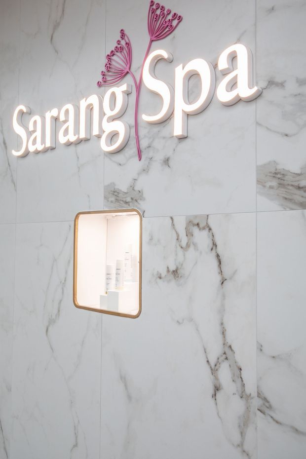 Close-up of a spa interior feature wall clad in large-format white marble-look panels with grey veining. A backlit “Sarang Spa” sign with a pink botanical motif is mounted above a small illuminated recessed display niche with a warm metallic frame.