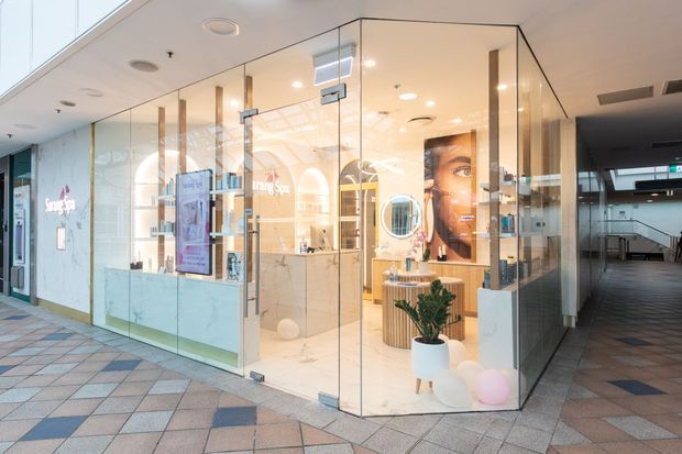 A bright contemporary beauty retail salon or commercial showroom enclosed by full-height frameless glass walls and doors. The interior features warm timber-toned cabinetry and display shelving, illuminated product displays, a reception or consultation counter, mirrors, potted greenery, and soft decorative lighting. The shopfront opens onto a tiled enclosed shopping-centre corridor, with a darker hallway visible to the right.