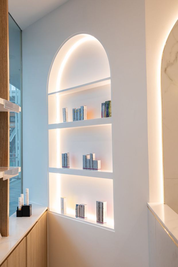 A refined built-in shelving niche with a tall arched top and integrated warm LED strip lighting. White painted shelving displays books, while pale timber cabinetry and a light stone or marble benchtop frame the lower portion. A curved illuminated architectural feature is partially visible on the right, with glazing and timber details at the left.