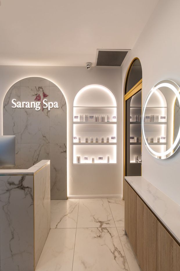 A refined spa reception and product display area featuring large white marble-look tiled flooring and wall cladding, an illuminated reception counter, arched recessed display shelving with integrated lighting, a round illuminated mirror, and warm timber cabinetry. Gold-toned trims and soft ambient lighting create a luxurious, contemporary commercial interior.