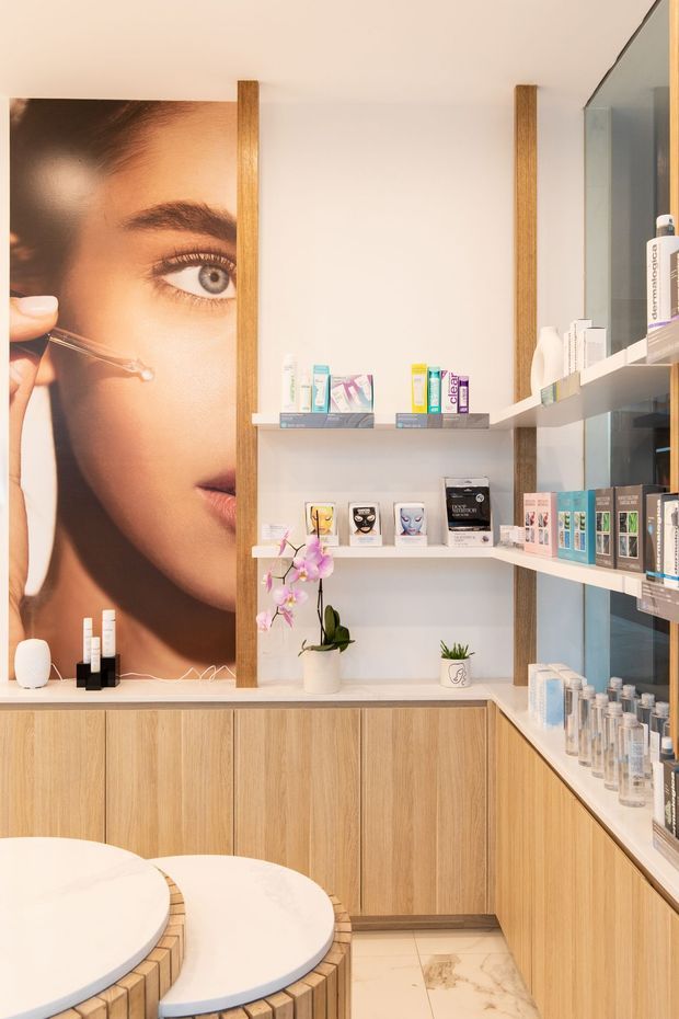 A bright, refined skincare retail or consultation space with light timber built-in cabinetry, open white display shelving, pale stone or marble-look countertops and floor tiles, and a large beauty photograph forming a feature wall. Shelves are stocked with skincare products, while a rounded white consultation table and curved timber seating occupy the foreground. A glass-fronted display area is visible on the right, with soft decorative orchid planting adding a subtle natural accent.