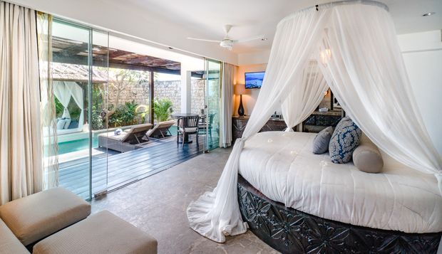 Bright, open-plan bedroom with a large four-poster bed draped in sheer white fabric, neutral upholstered seating, and full-height glazing opening to a timber deck and swimming pool courtyard. The room features polished stone-look flooring, white walls, extensive glass doors, and a relaxed resort-like atmosphere.