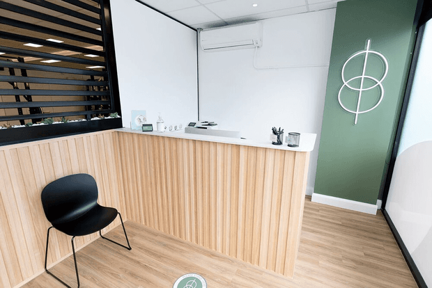 A bright, minimalist reception area with a light timber slatted reception desk, white benchtop, black visitor chair, and pale timber-look flooring. A dark green feature wall with a simple circular wall logo adds contrast, while black-framed horizontal timber screening separates the reception from the space behind it. The room has a clean commercial fit-out with a suspended ceiling and wall-mounted air conditioner.