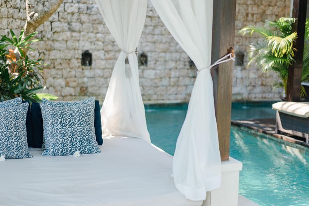 A bright tropical outdoor lounging area beside a turquoise swimming pool, featuring a white draped daybed with patterned cushions, timber posts, lush palms, and a rustic pale stone wall enclosure.