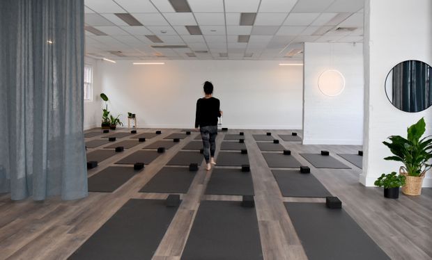 A spacious, minimalist yoga or fitness studio with rows of black exercise mats and small black yoga blocks arranged on a grey-brown timber-look floor. A person stands centrally in the studio. The room has white walls, a suspended acoustic tile ceiling with recessed lighting, a grey curtain partition, potted plants, and simple circular wall decor and mirrors.