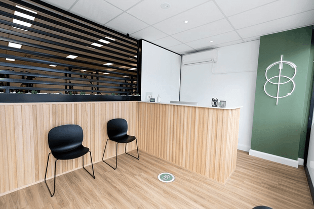 A modern commercial reception area with a long light timber reception desk and matching timber-look flooring. Two black visitor chairs sit in front of the desk. Behind the desk are white walls, a suspended acoustic tile ceiling, a wall-mounted air-conditioning unit, and a dark horizontal timber slat screen. A deep green feature wall with a simple white circular graphic provides contrast.