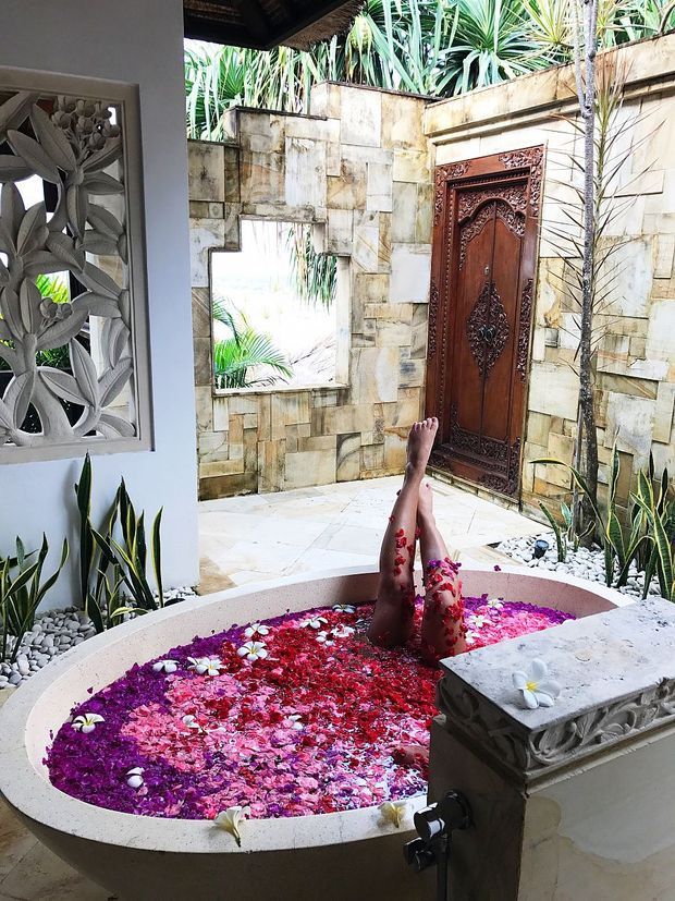 A tropical indoor-outdoor bathing area featuring a round sunken soaking tub filled with flower petals, a person bathing, pale stone paving and walls, lush tropical planting, an intricately carved dark timber door, and decorative carved stone detailing. The space has an exotic, resort-like character with strong ornamental craftsmanship.