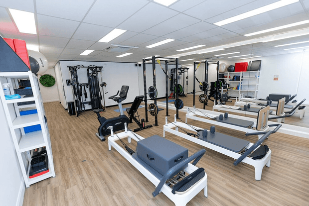 Bright, spacious commercial Pilates and fitness studio with multiple reformer machines, wall-length mirrors, cable and strength-training equipment, open shelving, and light wood-look flooring beneath a suspended grid ceiling with fluorescent panel lighting.