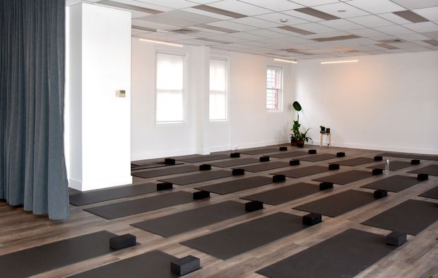 A bright, spacious studio room arranged for group yoga or fitness classes, with numerous dark exercise mats and small yoga blocks laid out in orderly rows on a wood-look floor. The room has white walls, tall windows with blinds, a suspended acoustic tile ceiling, a large dark curtain at the entrance, and minimal plant and storage elements.