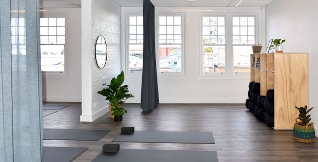 Bright, open yoga or fitness studio with white-painted brick walls, large multi-pane windows, dark timber-look flooring, grey exercise mats, and a dark curtain divider. Potted plants and simple timber shelving add natural accents to the clean, airy space.