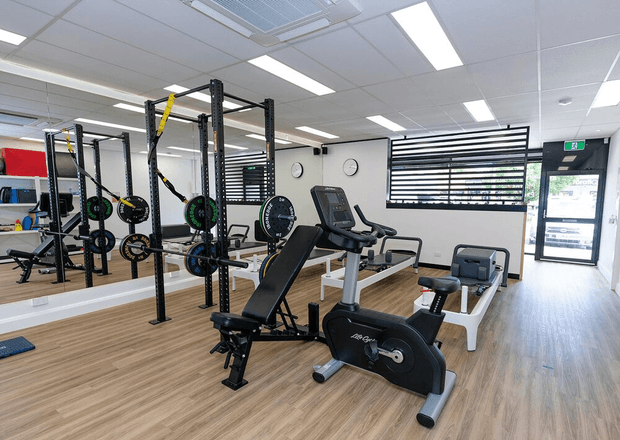 A bright, modern commercial fitness studio with black strength-training racks, weight plates, benches, treadmills, and stationary exercise bikes. The room has a light timber-look floor, white walls, mirrored surfaces, horizontal black window blinds, and a suspended acoustic ceiling with linear fluorescent lighting. A glazed entrance door and green emergency exit sign are visible at the rear.