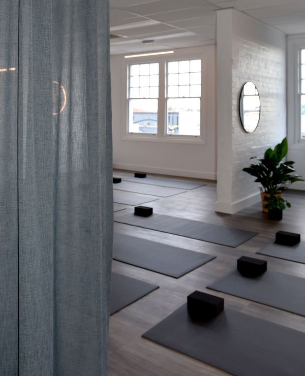A bright, minimalist yoga or fitness studio with rows of grey exercise mats and black yoga blocks arranged across a timber-look floor. The space features white-painted brick walls, large divided-pane windows, a simple suspended ceiling, a round wall mirror, and a potted plant. A translucent grey curtain partially frames the view in the foreground.