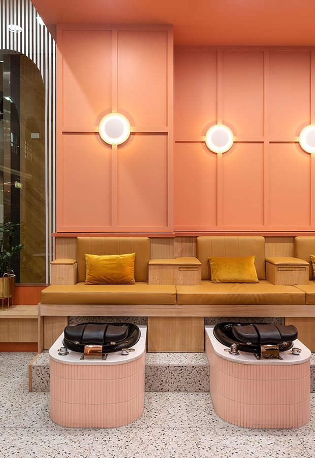 A warmly lit, highly stylised commercial salon interior featuring two pedicure stations with black foot basins set into rounded blush-pink bases. A long caramel leather banquette runs along a peach-toned wall with geometric applied panelling and circular glowing wall lights. Terrazzo flooring and stepped terrazzo platforms add texture, while timber-toned joinery and vertical slatted screening appear at the left.