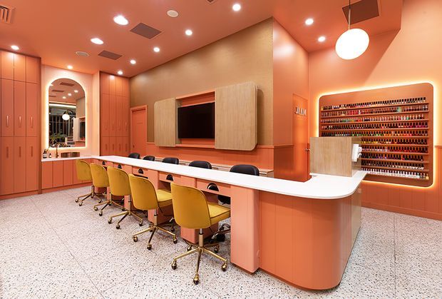 A brightly lit contemporary commercial beauty salon or nail studio with a long curved manicure counter and multiple mustard-yellow rolling chairs. The space features warm coral and terracotta tones, integrated cabinetry, a wall-mounted television with timber surround, an illuminated product display shelving unit, terrazzo flooring, recessed ceiling lights, and a large round pendant light. An arched mirror and additional workstations are visible at the rear.