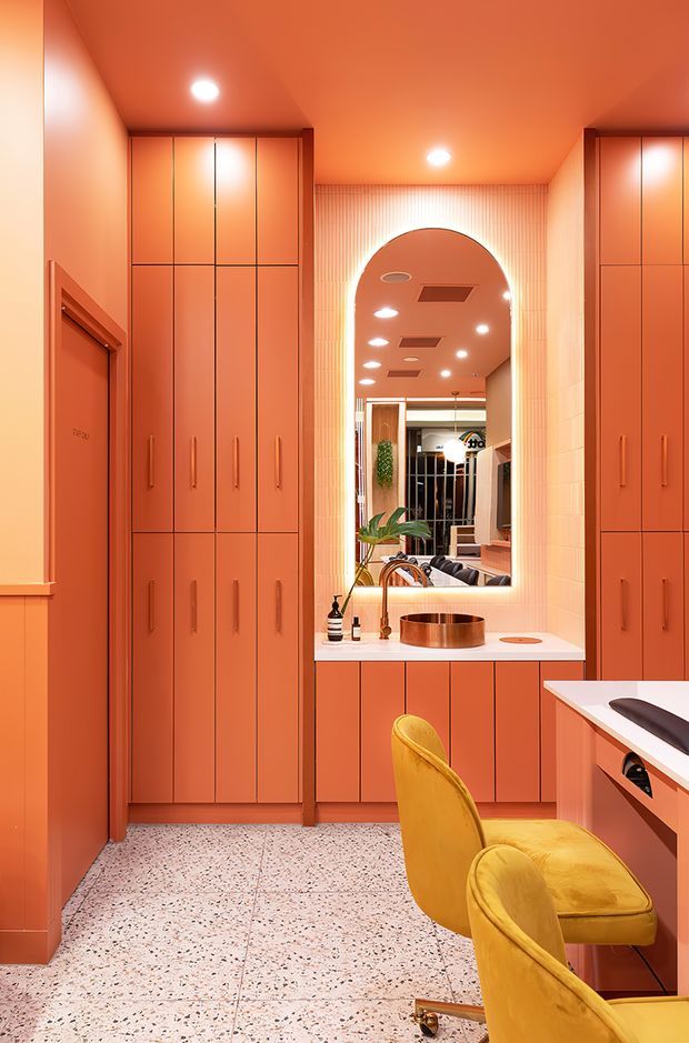 A vivid coral-orange dressing room or walk-in wardrobe with floor-to-ceiling built-in cabinetry, long vertical handles, a white benchtop vanity, and a large illuminated arched mirror. Two mustard-yellow upholstered chairs sit at the vanity. The room has a terrazzo floor, recessed ceiling lights, and a highly saturated, playful contemporary fit-out.