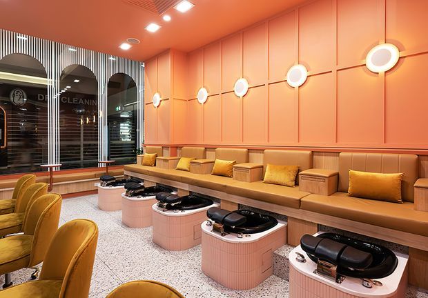 A stylish commercial salon interior with a warm coral-pink palette. The room features a long built-in upholstered bench with mustard cushions, individual pink tiled manicure or pedicure stations with black equipment, curved mustard lounge chairs, terrazzo-style flooring, decorative wall panelling, and illuminated circular wall lights. Arched glazed openings and a striped screen add visual interest.