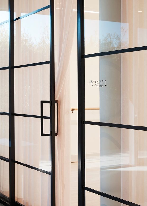 Close-up architectural photograph of a black metal-framed glazed door or partition with rectangular grid divisions, a bold rectangular pull handle, and sheer white curtains. A softly lit pale interior with timber handrail or trim is visible beyond the glazing, creating a refined, minimal composition.