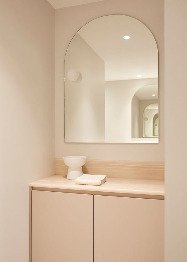 A softly lit minimalist bathroom vanity in warm beige tones, featuring a floating two-door vanity with a pale timber-look benchtop and backsplash, an arched wall mirror, integrated recessed ceiling lights, and a small basin with folded white towel. Repeated arched reflections create depth and rhythm.
