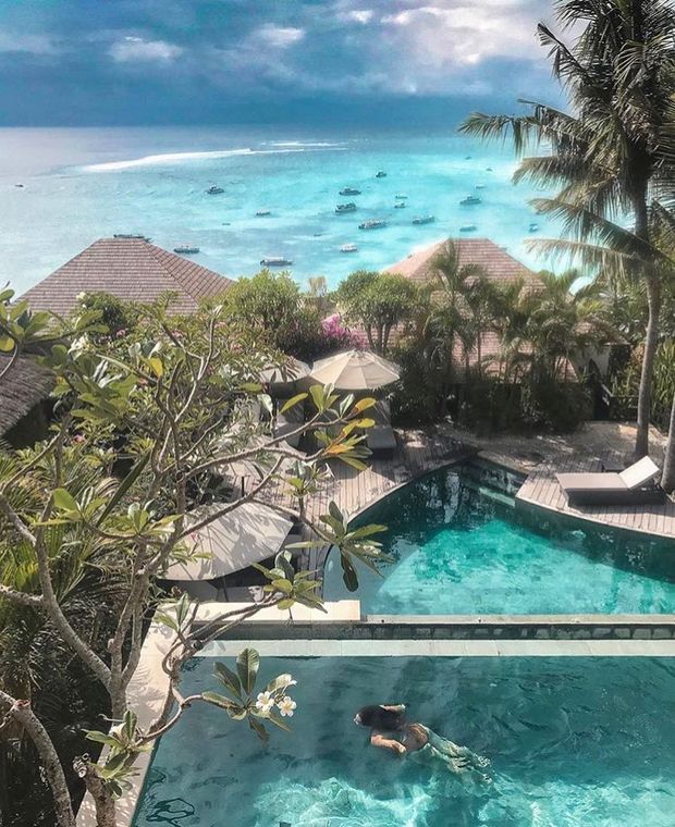 A tropical resort-style pool area overlooks a vivid turquoise ocean dotted with boats. The scene includes multiple tiered swimming pools, timber pool decking, sun loungers and umbrellas, lush palms and flowering tropical planting, and red-tiled pavilion roofs beneath a partly cloudy sky.