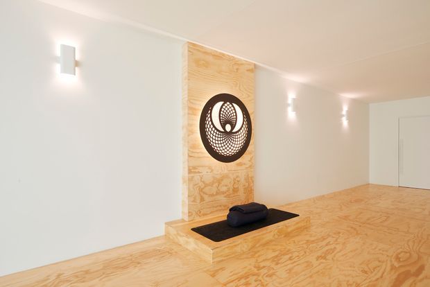 Minimalist interior hall or entry space with pale plywood flooring and a raised plywood platform featuring a black inset surface and circular sculptural artwork. White walls, concealed doors, and simple wall-mounted sconces create a clean, gallery-like atmosphere.