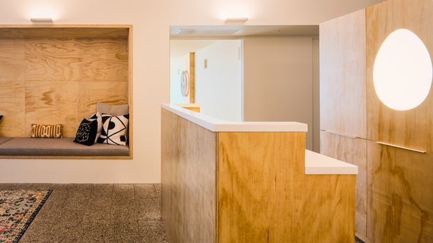 Warm contemporary entry area featuring a built-in timber-lined window seat with cushions, a patterned rug, and a sculptural timber reception or stair-side joinery element with white benchtop surfaces and an integrated circular wall light. A doorway and framed artwork are visible beyond.