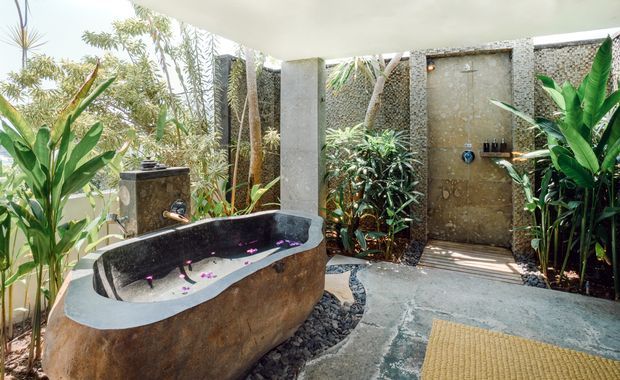 Open-air tropical bathroom with a freestanding stone bathtub, wall-mounted shower, concrete finishes, pebble landscaping, and dense lush planting surrounding the space.