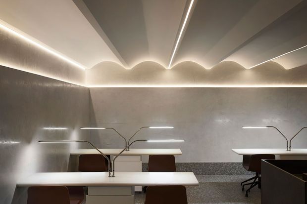 A sleek contemporary workspace with several white desks, sculptural minimalist desk lamps, and dark upholstered office chairs. The room features warm concealed LED lighting, a textured grey wall finish, a sculpted wave-like ceiling detail, and a speckled lower wall or dado surface. The composition creates a refined, atmospheric shared office environment.