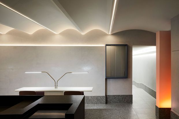 A refined contemporary interior featuring a dark stone or terrazzo dining table in the foreground, a slim white counter with sculptural brass-toned lighting, textured grey wall finishes, integrated linear LED lighting, and a softly scalloped ceiling detail. A dark-framed built-in cabinet and illuminated corridor enhance the minimalist, gallery-like composition, with a warm terracotta-orange vertical feature adding contrast.